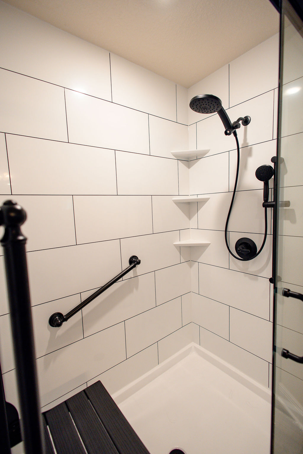 LatashaCarrawayPhotographyAFTER-8662 detail shot of walk-in shower accessories, including detachable shower wand, corner shelves, grab bar, and fold down black wooden seat
